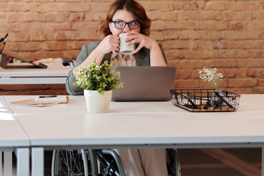 A woman in a wheelchair drinks coffe at a desk. It looks like she has something on her mind.