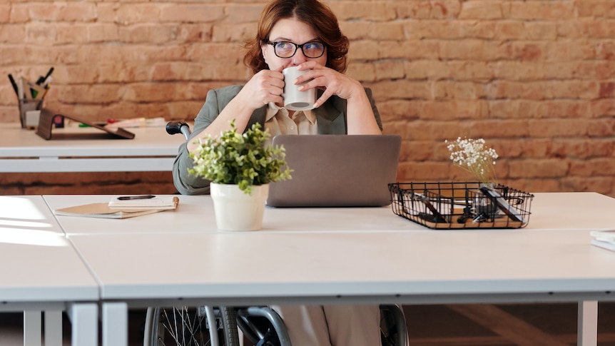 A woman in a wheelchair drinks coffe at a desk. It looks like she has something on her mind.