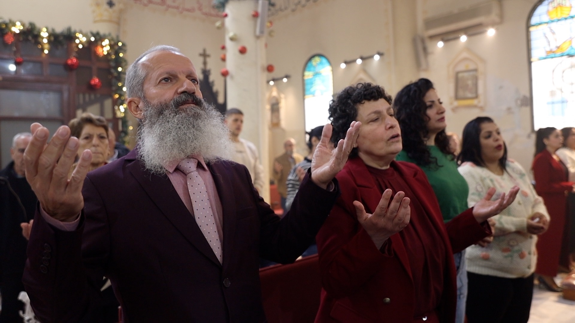 Un hombre con una larga barba blanca y traje reza en una iglesia decorada con los brazos abiertos y las palmas hacia arriba.