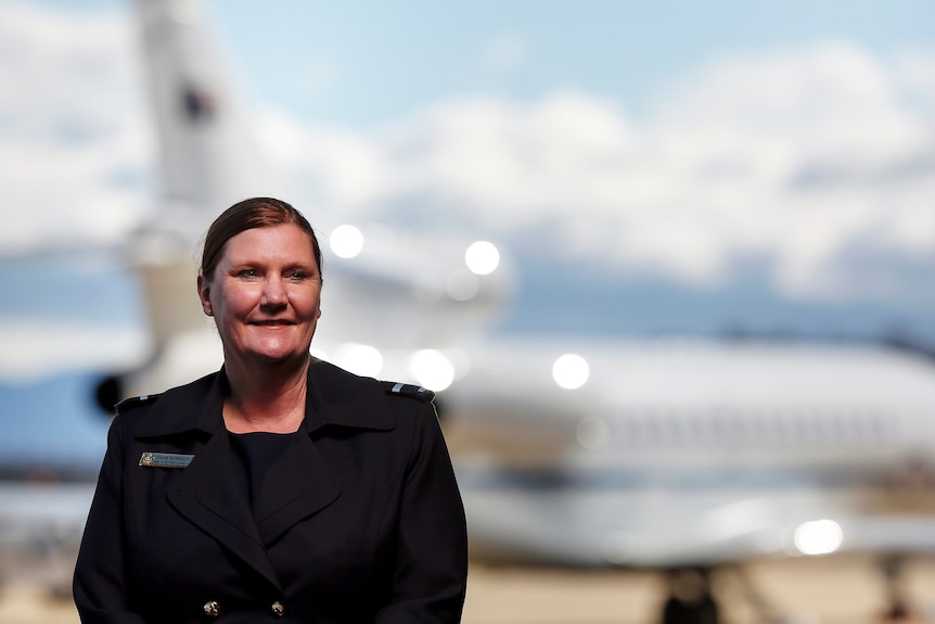 A woman wearing a black blazer with her hair back in a low bun standing in front of an out of focus airplane