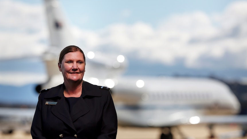 A woman wearing a black blazer with her hair back in a low bun standing in front of an out of focus airplane