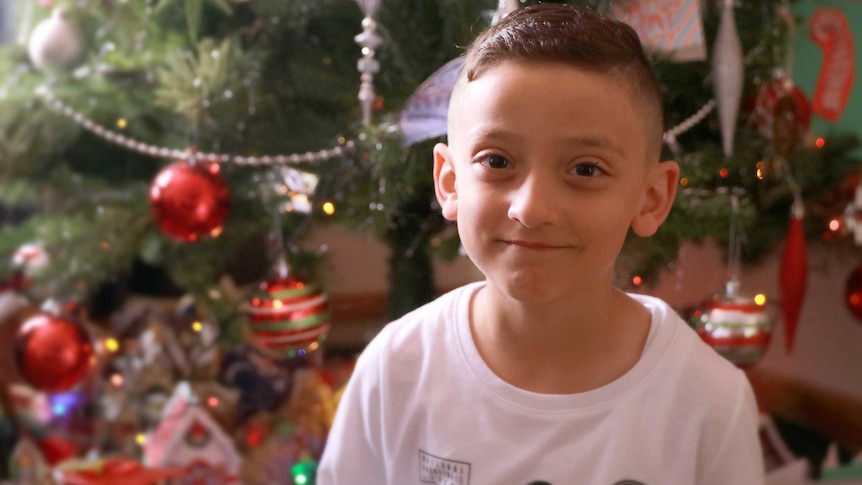 A young boy smiles in front of a Christmas tree.