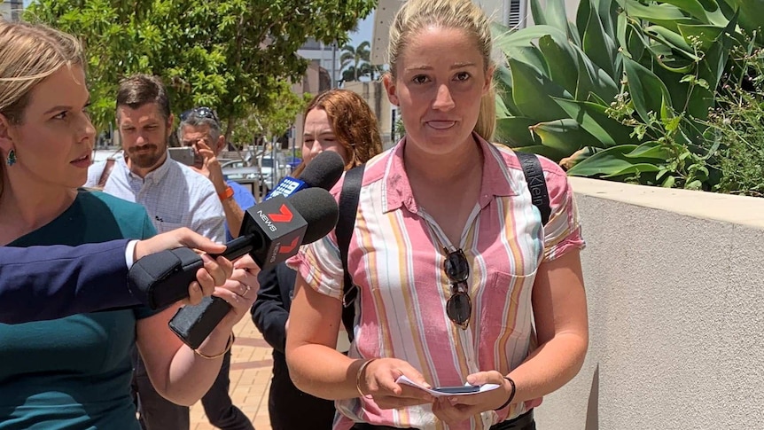A young blonde woman walks away from a court building, flanked by journalists.