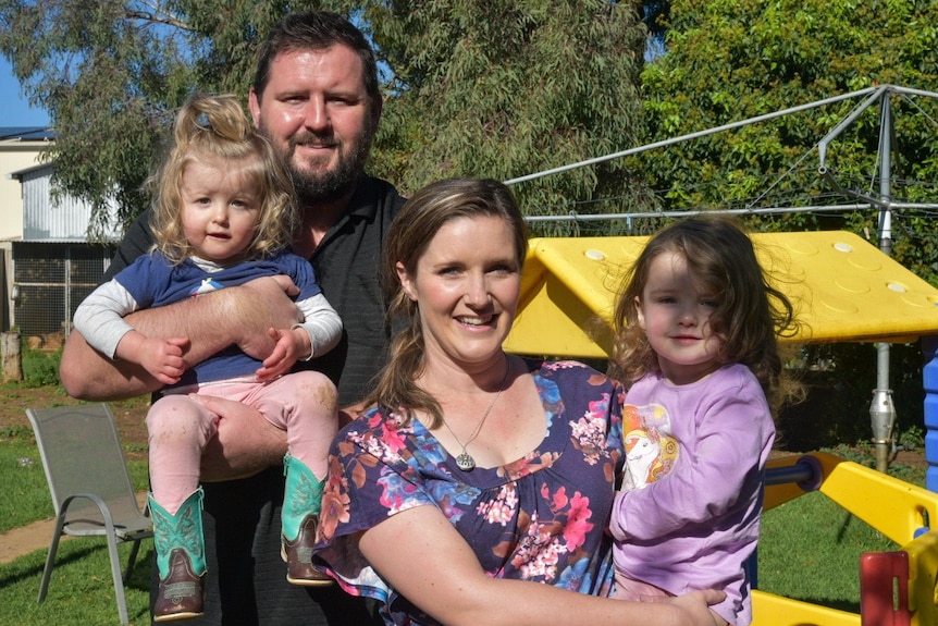 A man and a woman each holding a young girl in front of a child's plastic play house, and a clothes line.