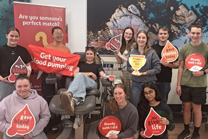 A group of young people holding signs of blood donation while one in a chair donates blood