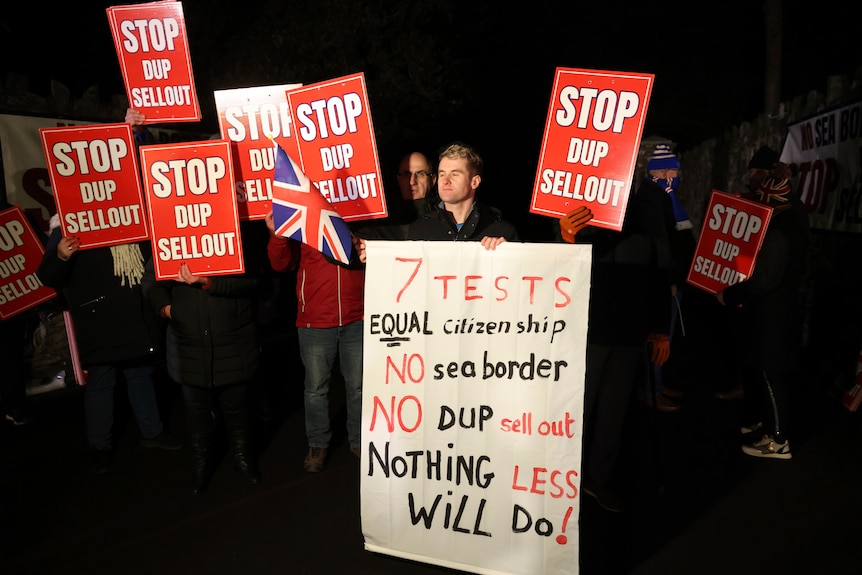 Protesters gathered at night carry banners reading 'Stop DUP sellout', one carrying a Union Jack flag