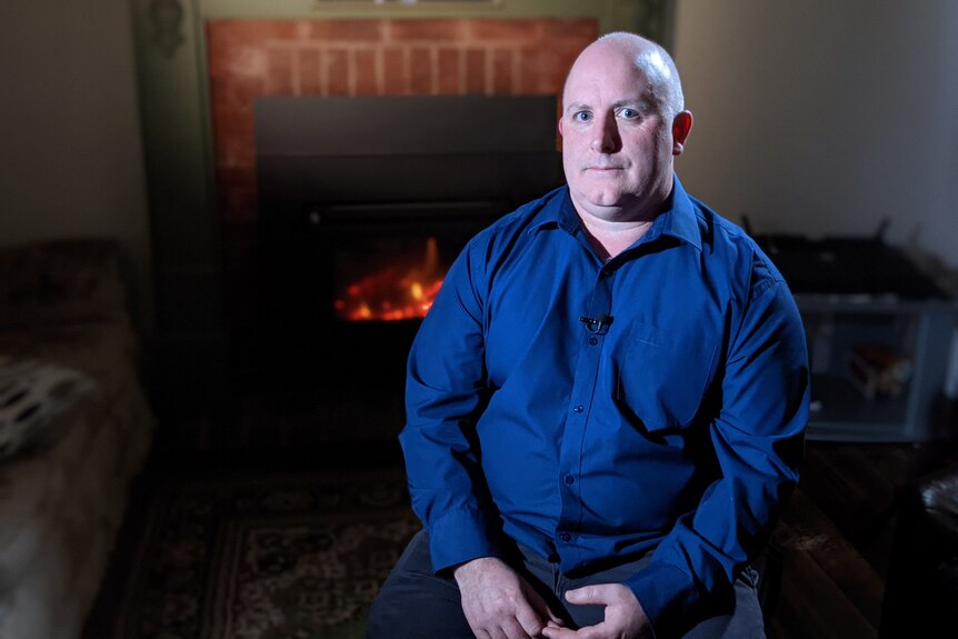 Man wearing blue shirt sitting in front of a fireplace.