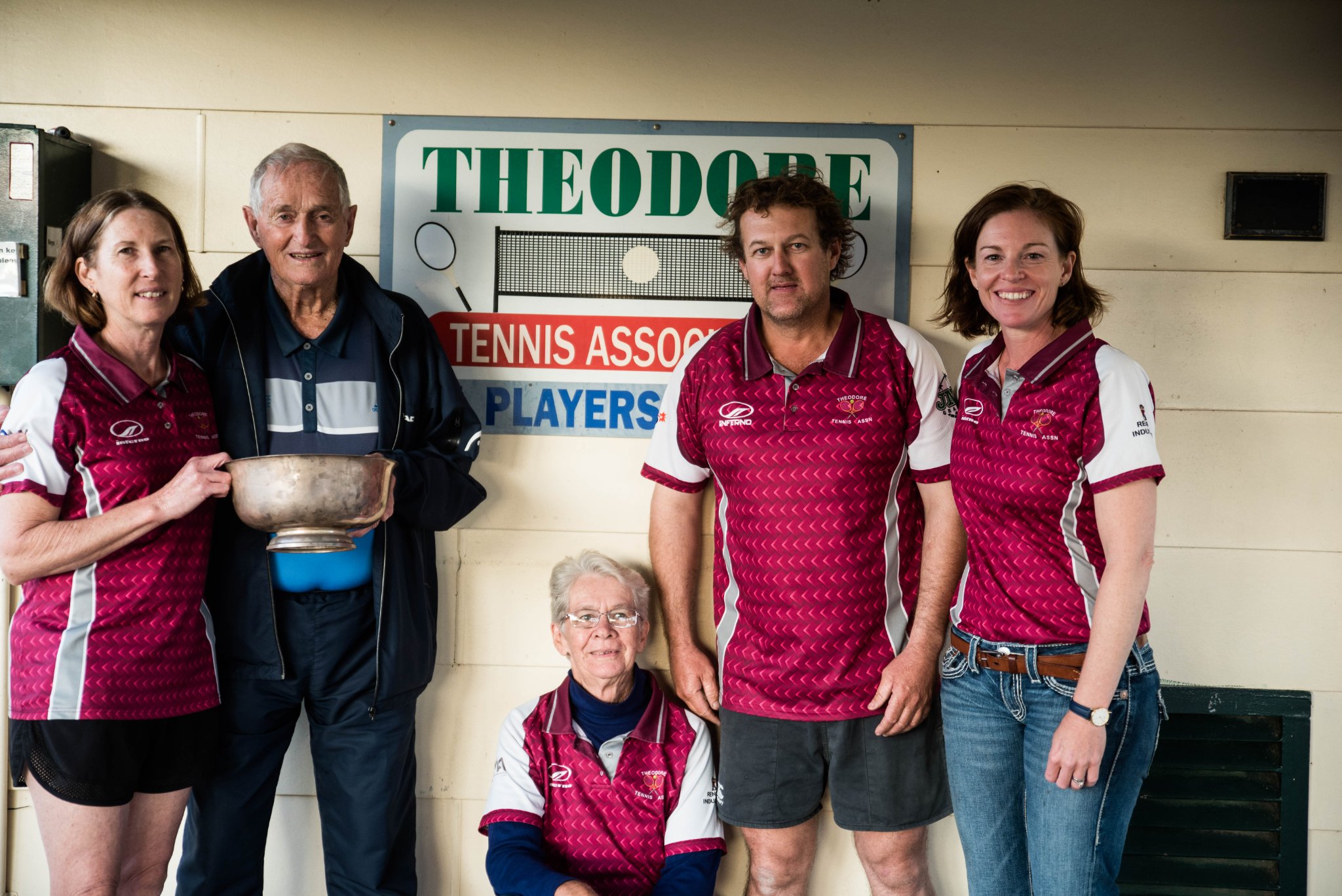 A man with an old trophy with a group of people in front of a sign for the Theodore Tennis Association