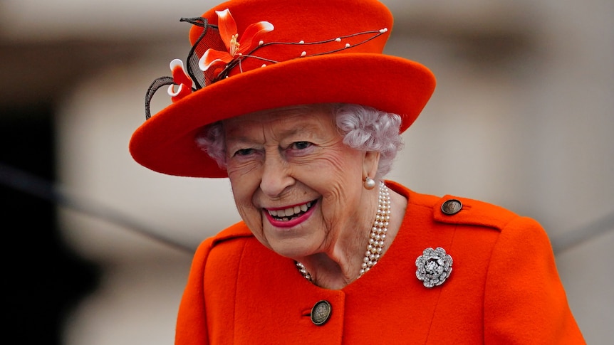 Queen in red blazer and hat while smiling.