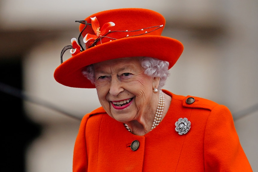 Queen in red blazer and hat while smiling.