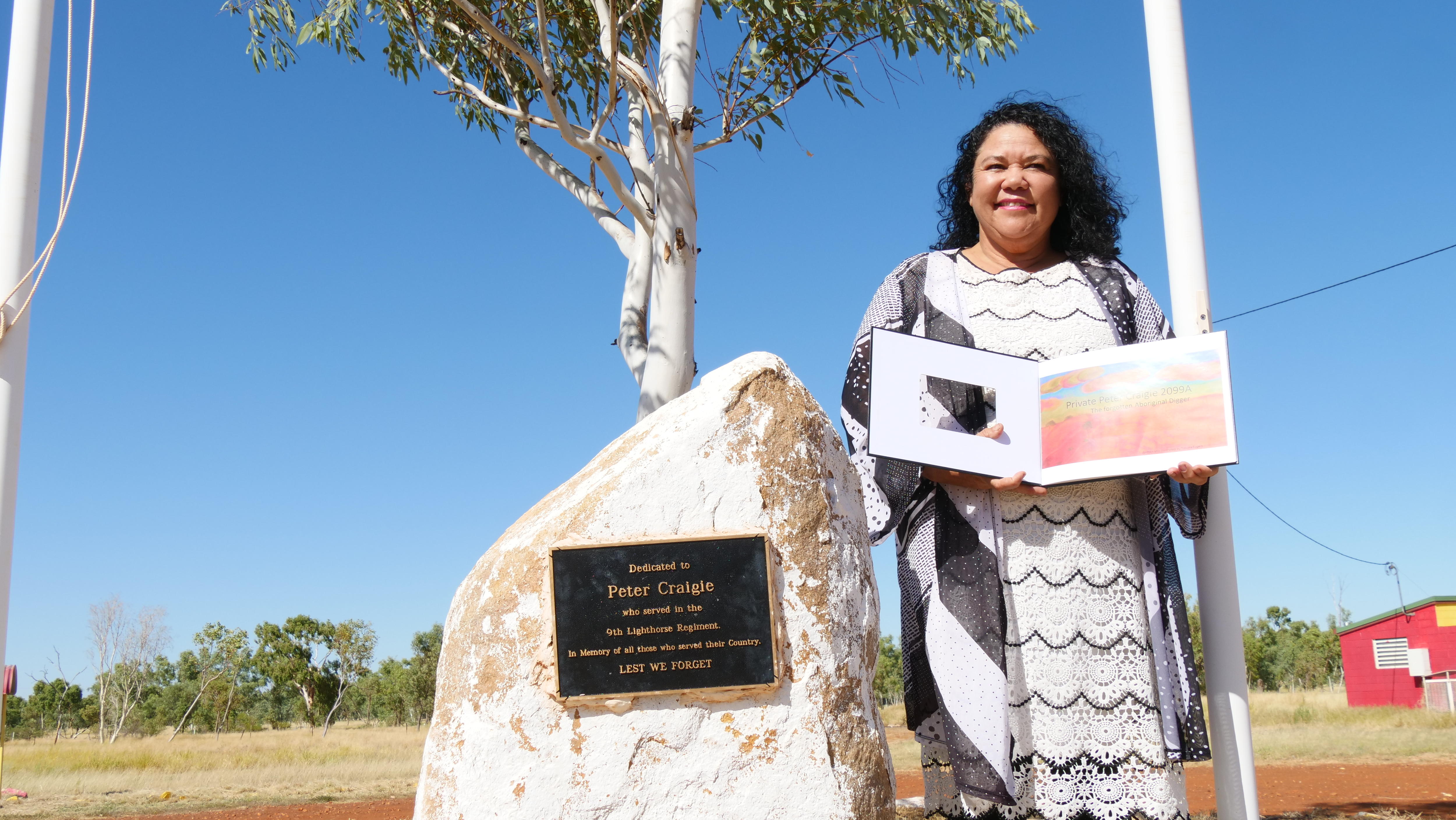 Woman stands next to white stone war memorial holding an open picture book.