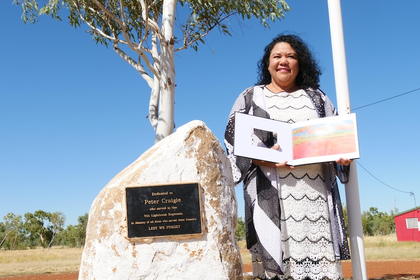 Woman stands next to white stone war memorial holding an open picture book