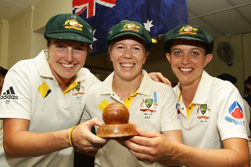 Three Australian women cricketers hold a cricket ball trophy and smile at the camera.