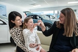 A businesswoman sits in a car dealership with her young daughter on her lap. A female colleague holds up keys to the daughter.
