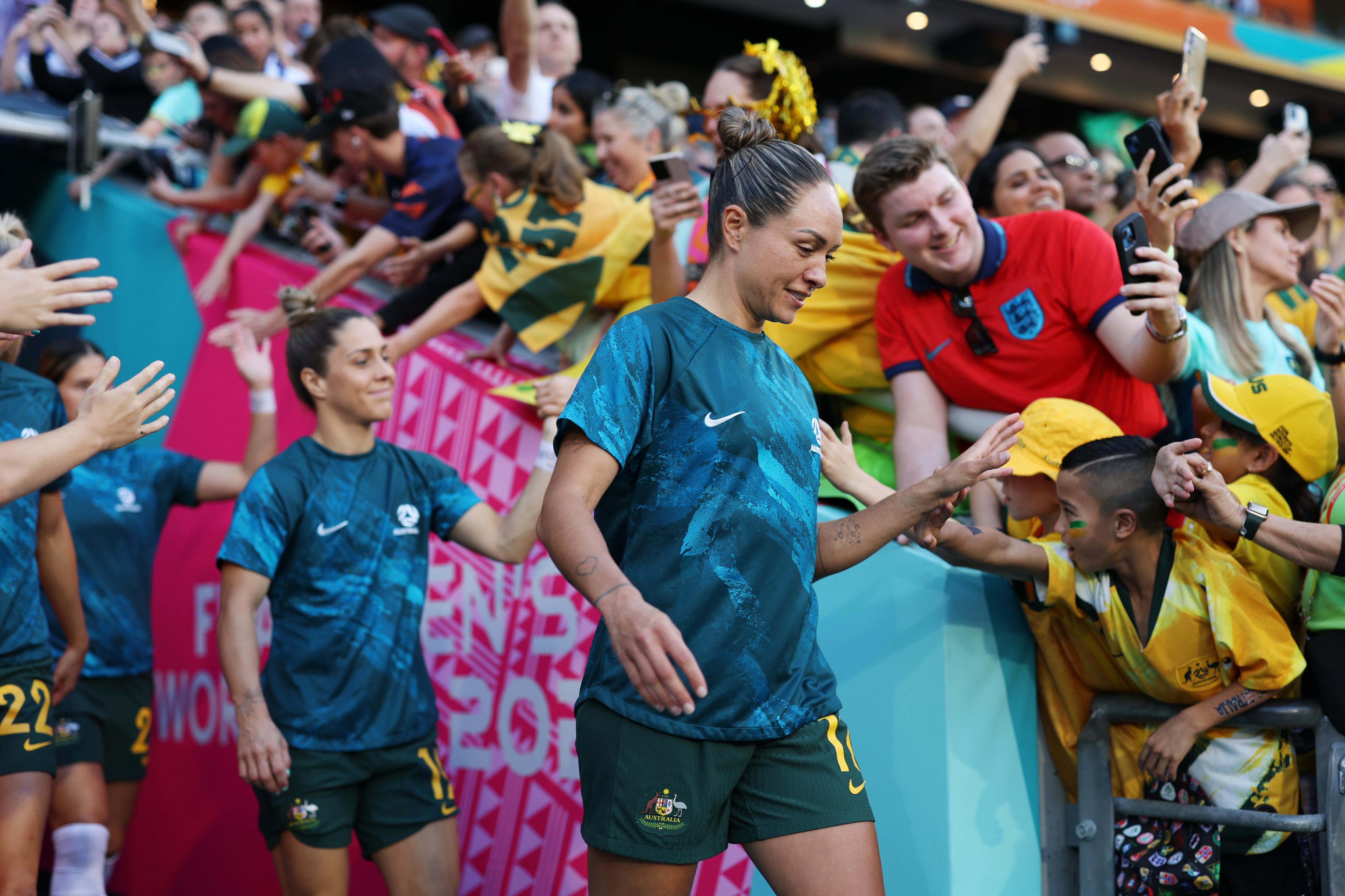 Dos jugadores de fútbol con camisas azules y pantalones cortos verdes salen de un túnel con fanáticos con amarillo detrás de ellas