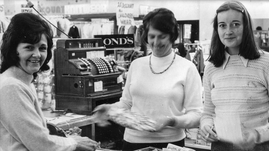 Three women counting money at a cash register inside a department store.