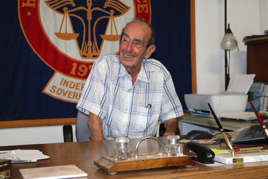 Prince Leonard smiles while sitting at a desk in front of a large blue, red and white flag.