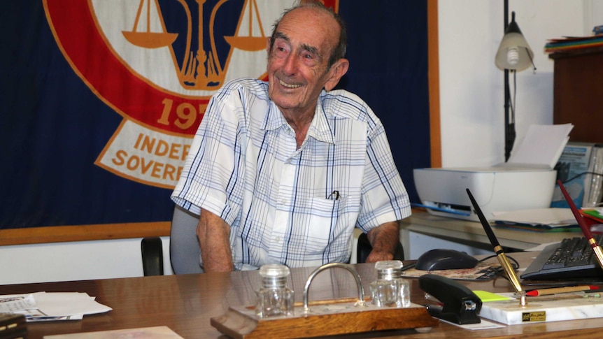 Prince Leonard smiles while sitting at a desk in front of a large blue, red and white flag.