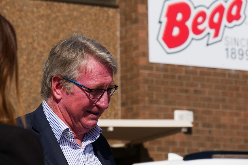 A bespectacled middle-aged man in a suit standing near a sign that says "Bega".