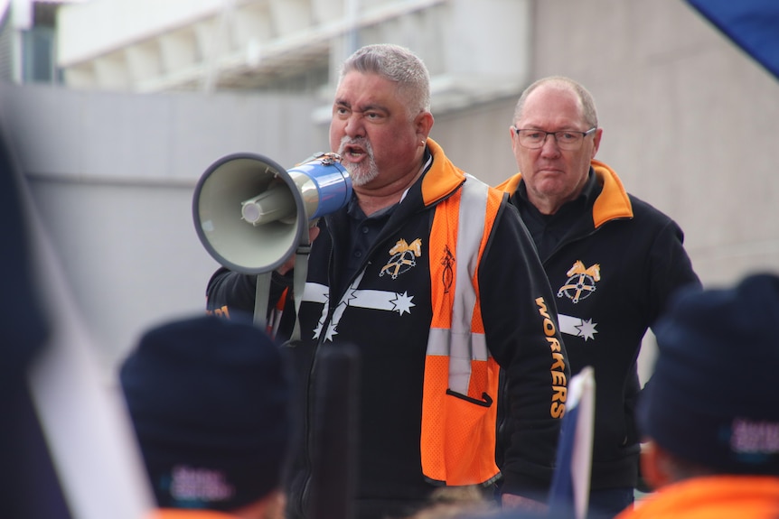 A man talks into a megaphone and a man stands behind him on a stage