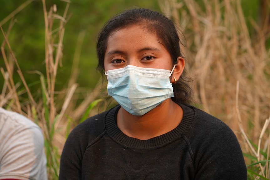 A teenage girl with dark hair wears a medical mask.