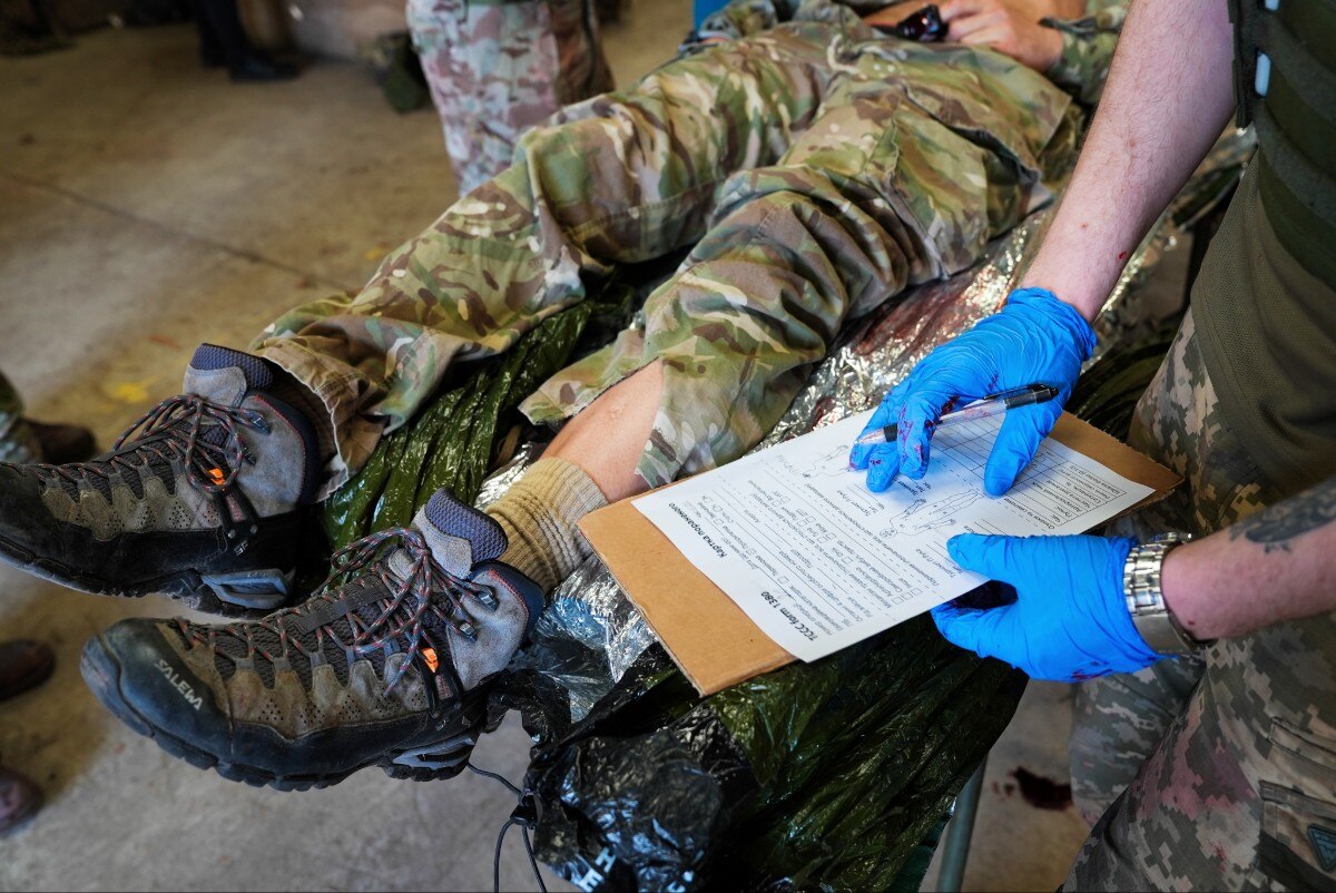 A person with a clipboard standing over another person who's lying down. Their faces are not visible. They are both in uniform