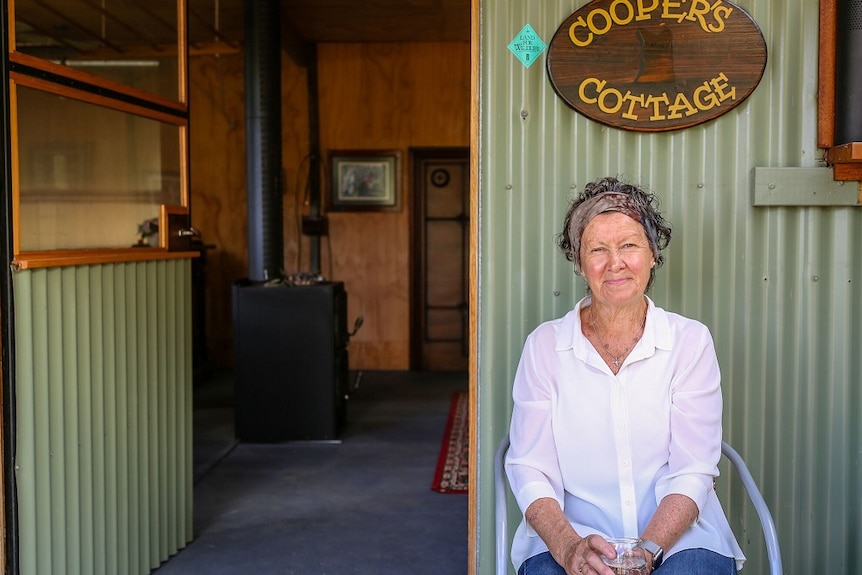 A woman in a white shirt sits in front of a corrugated green tin shed with a fireplace inside.