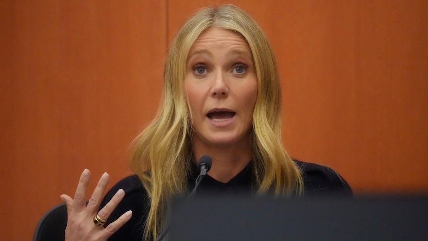 A blonde woman is pictured speaking into a microphone as she gestures with one hand.