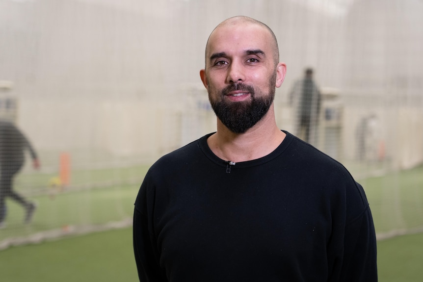 A bald man with a beard dressed in black poses at an indoor cricket centre.