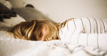 Woman lying face down on bed with her hair obstructing her face and looking tired to depict sleep problems.