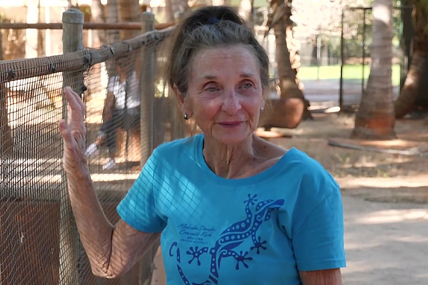 An older woman in  a blue t-shirt stands next to a wire fence.