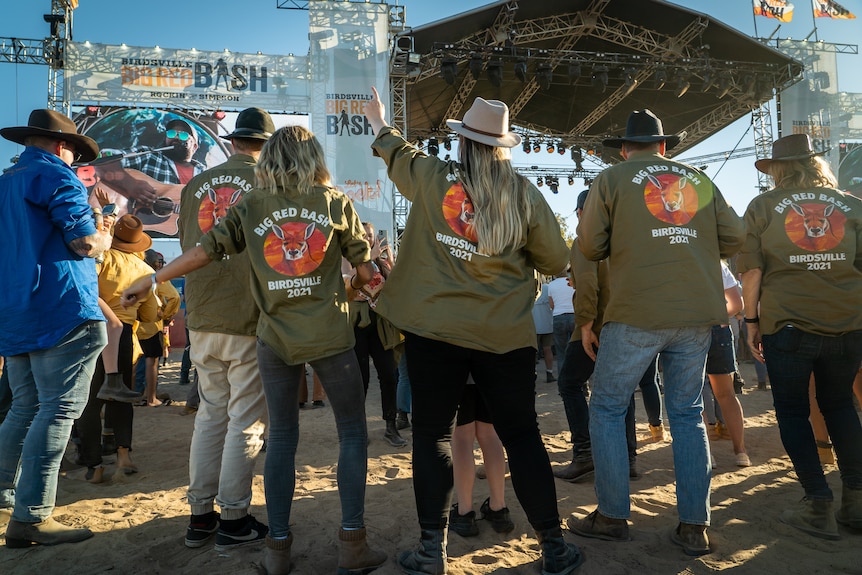 People wearing kahki green 'Big Bash' shirts with their arms up dancing to a concert.