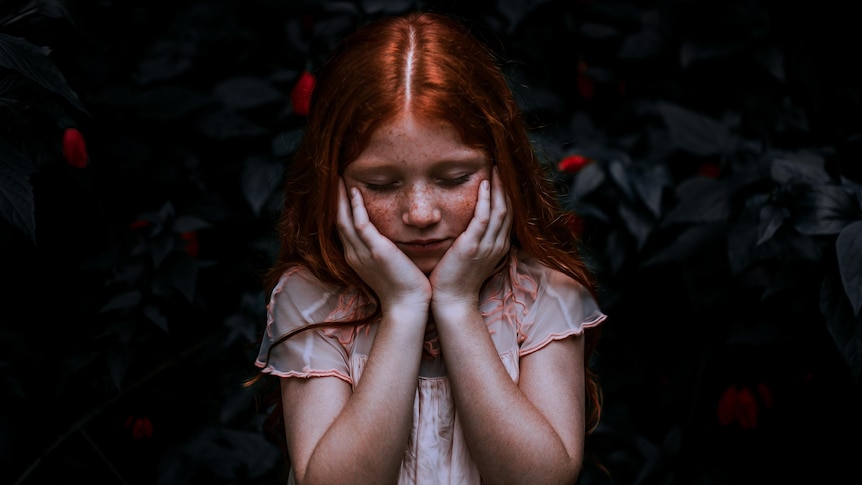A moody photo of a pale young girl with red hair holding her head in her hands
