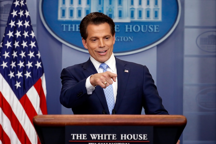 A man in a suit stands at a lectern with an American flag, pointing