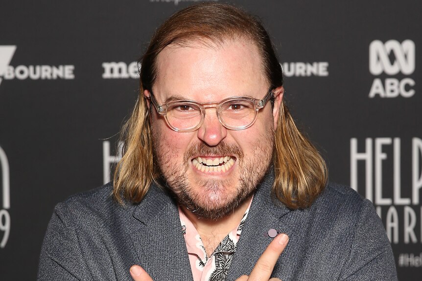 Brent Hill with shoulder-length blond hair and glasses and in grey suit, posing on red carpet of Helpmann Awards.