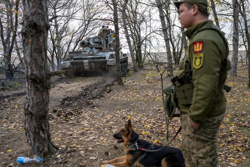 A Ukrainian fighter stand with a German Sheppard dog on a leash near a tank.