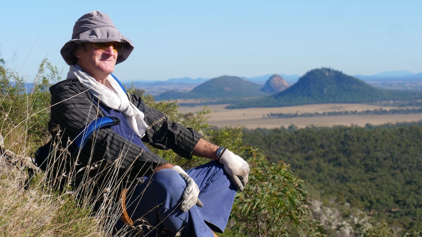 Man sitting with his legs crossed wearing a hat, sunglasses, gloves and long pants. Mountains behind.