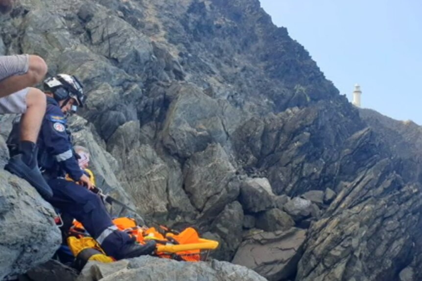 A rocky cliff with a lighthouse in the background and paramedic to the left of shot, with orange, hi-vis rescue apparatus 
