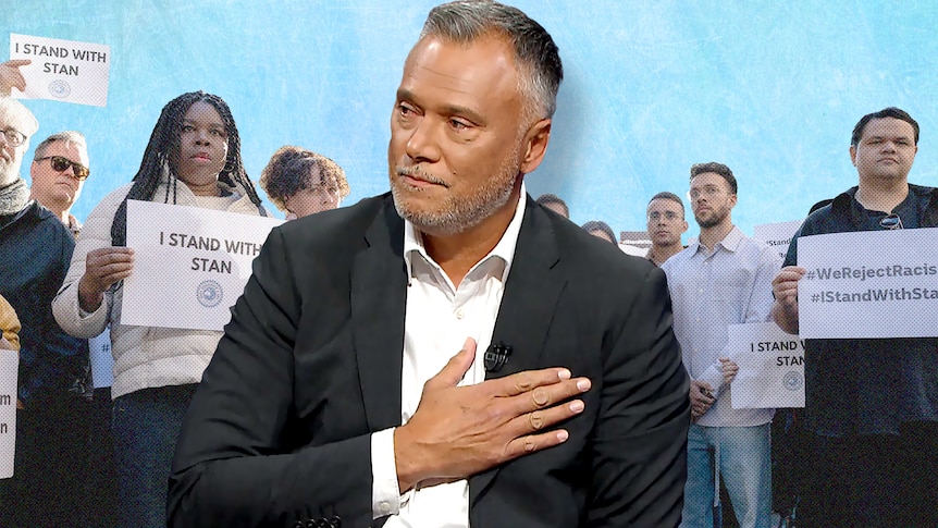 Stan Grant places his hand over his heart. ABC staff at a rally with pacquards saying I stand with Stan in the background.