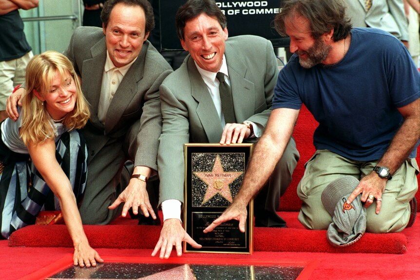 A man wearing a tie is surrounded by three people kneeling on a red carpet in front of a pink marble star.