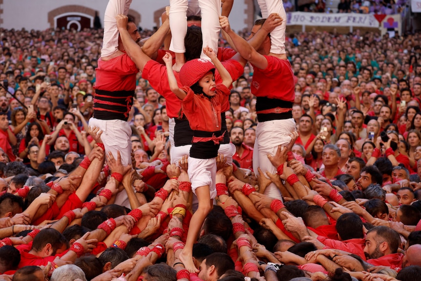 A small child wearing a red shirt and helmet stands on top of a mass of people in red, arms raised in celebration.