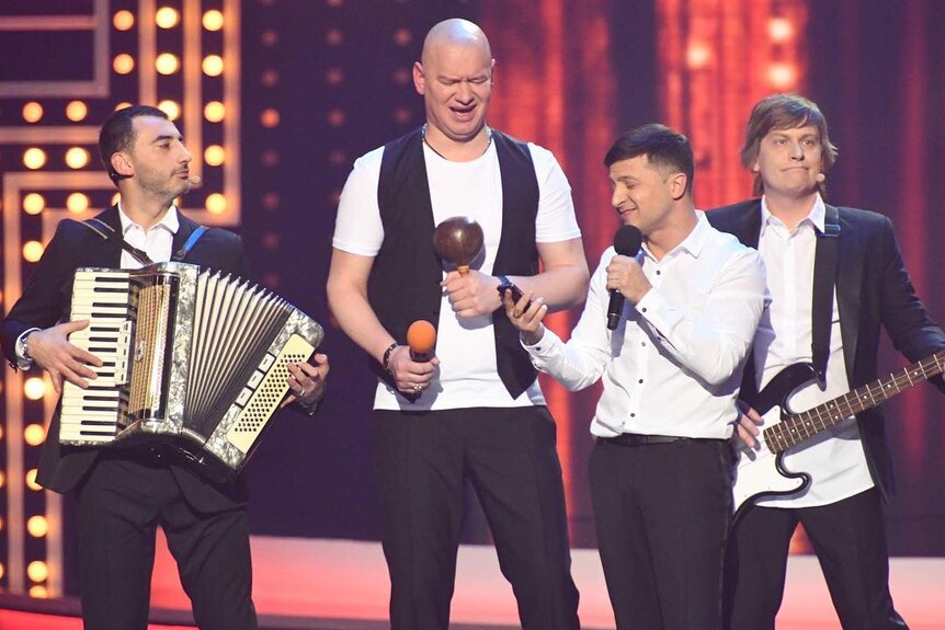 Volodymyr Zelenskyy on stage with four other men holding instruments 