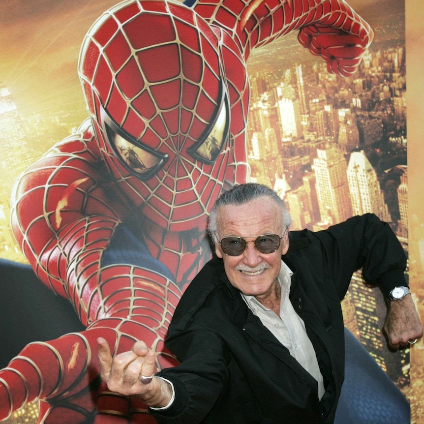 A man wearing dark glasses adopts a Spider-Man pose in front of a Spider-Man poster