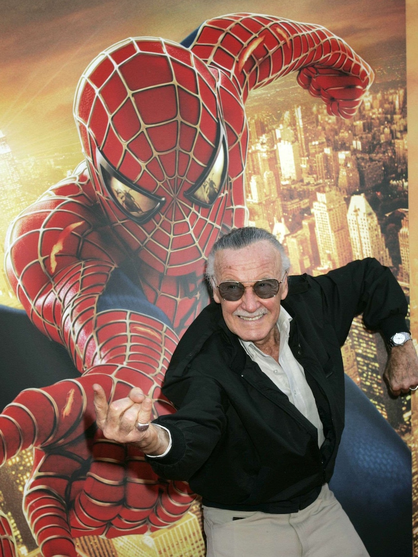 A man wearing dark glasses adopts a Spider-Man pose in front of a Spider-Man poster