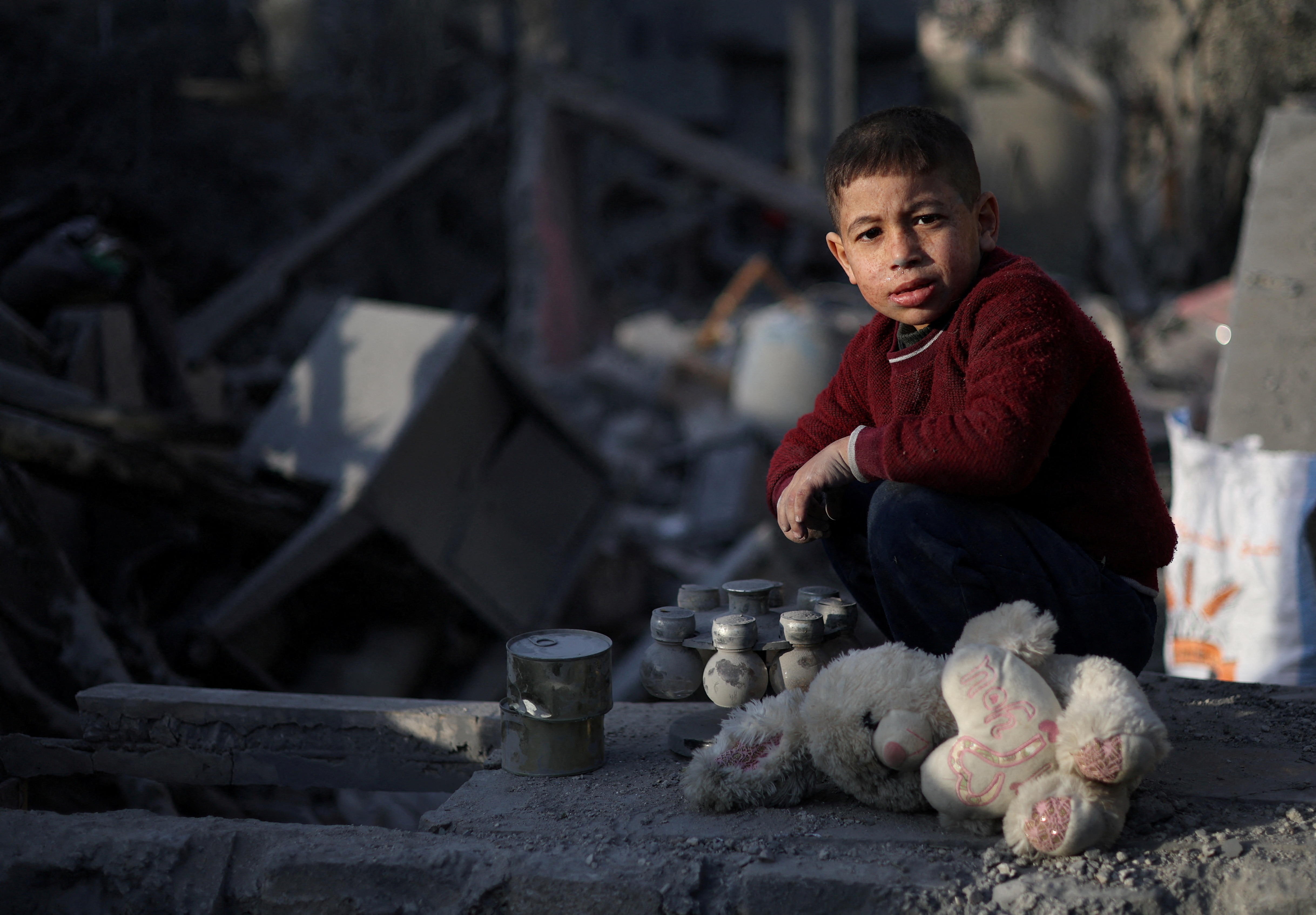 A little boy in a red jumper kneels by wreckage and a teddy