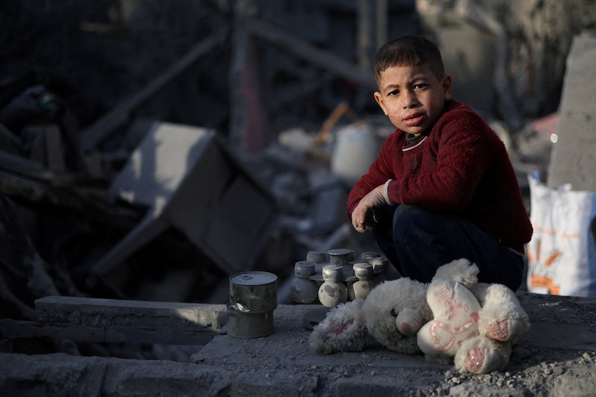 A little boy in a red jumper kneels by wreckage and a teddy