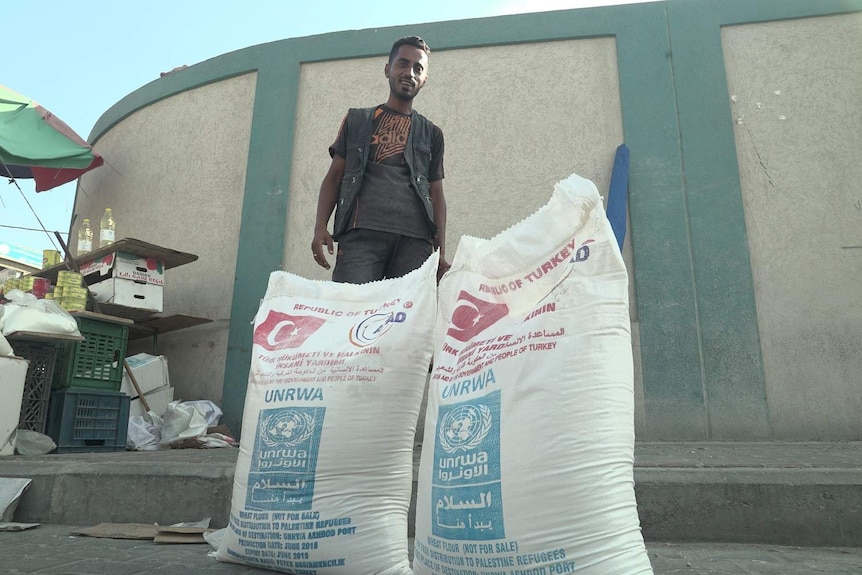 A man stands behind two large sacks of flour with the imprint of UNRWA.