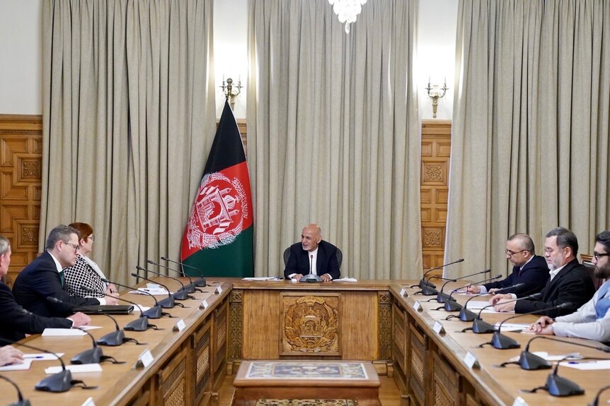People sit in a formal meeting room with an Afghan flag in the background. 