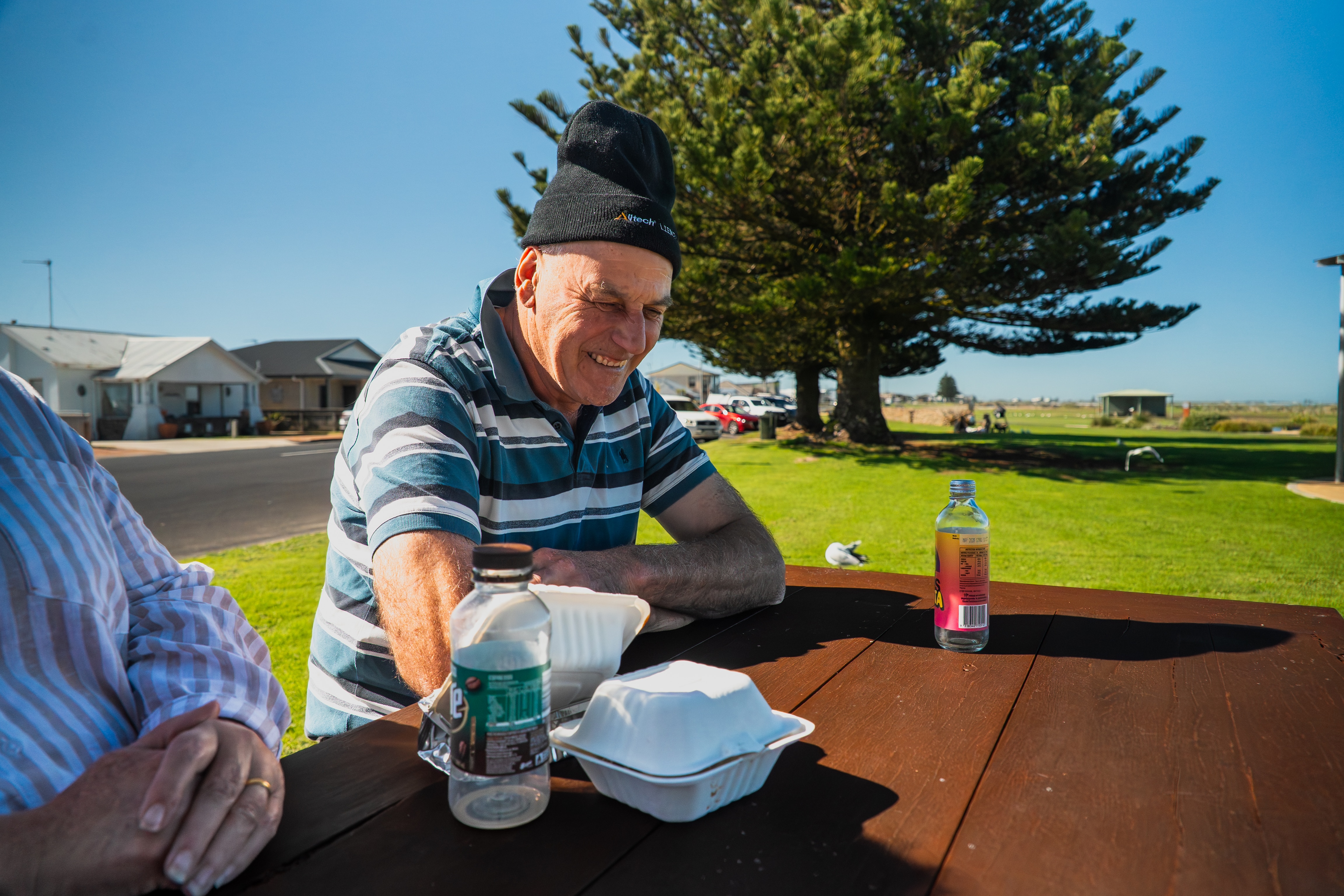 A smiling man sits on park table after eating lunch; seagulls can be seen on the freshly mowed grass behind him.
