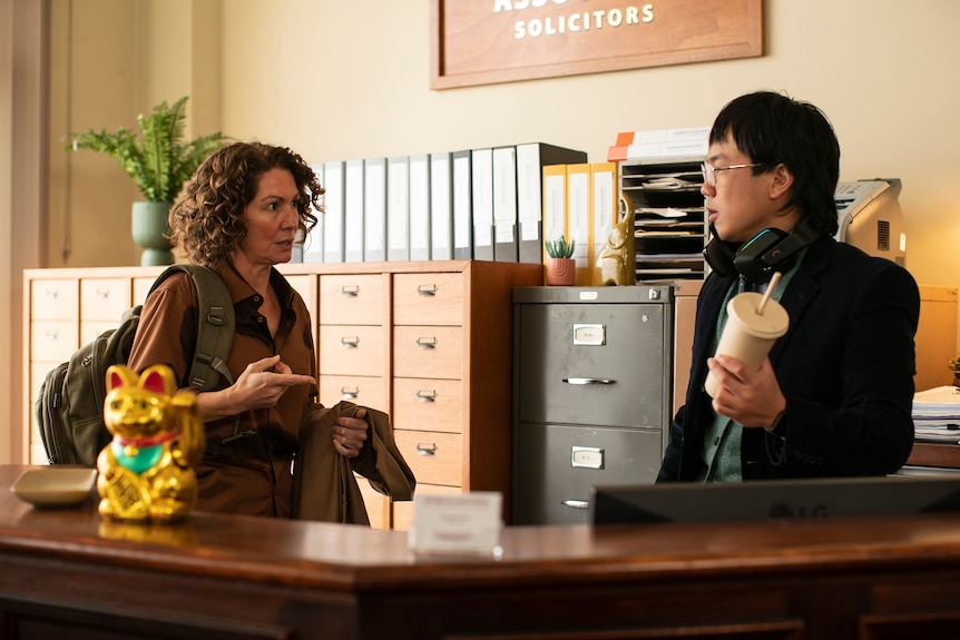 演员 Kitty Flanagan 和 Aaron Chen 在 ABC 电视台的 Fisk 喜剧中的一个场景中在办公室聊天。